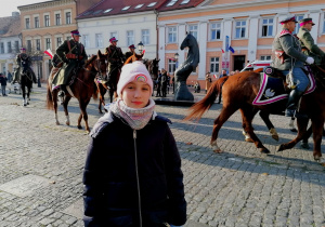 Dziewczynka stoi na Placu Wolności w Koninie, a za nią na koniach przechodzą żołnierze.